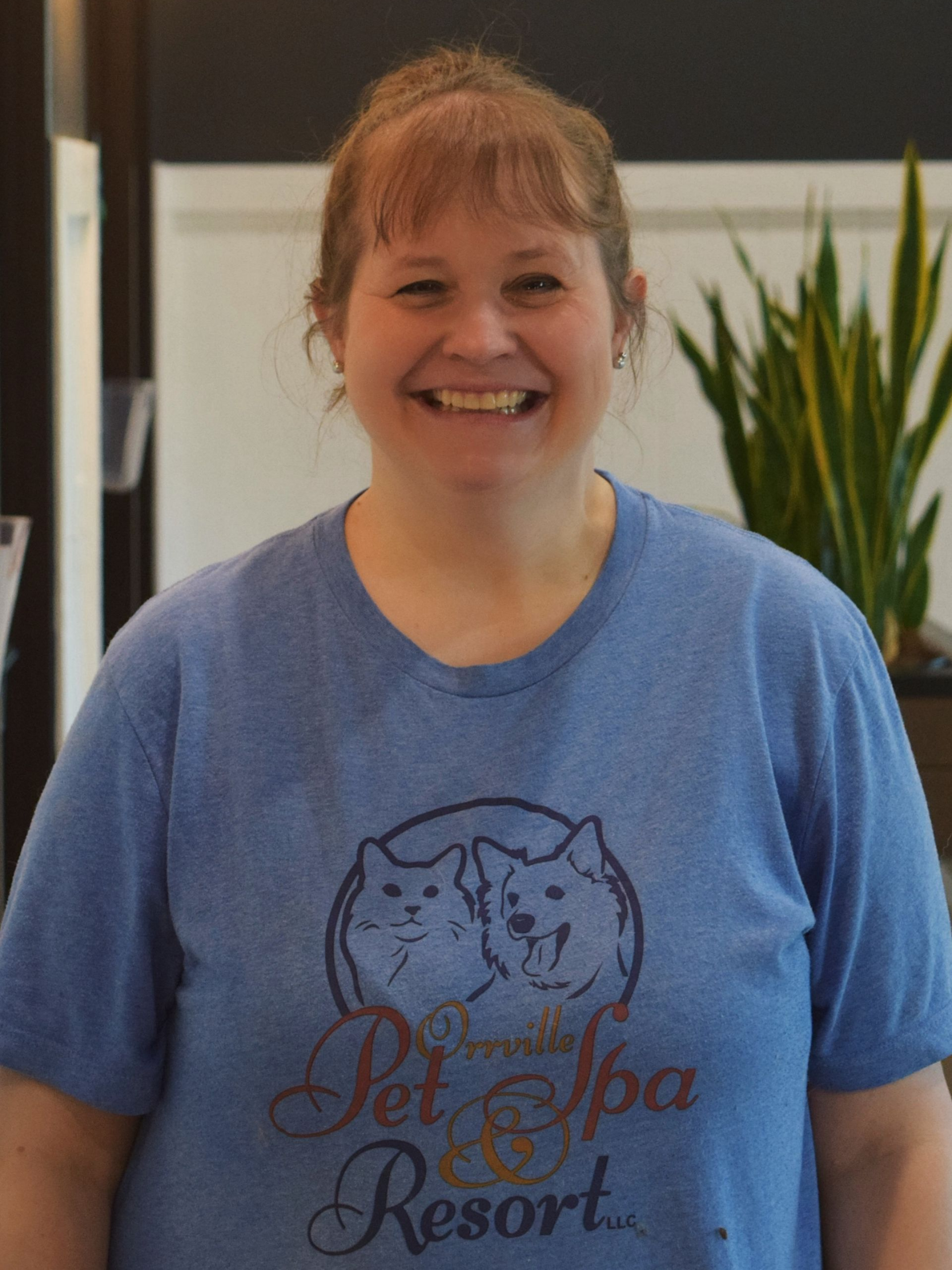 Woman with short brown hair smiles, wearing a blue t-shirt with a small logo, indoors.