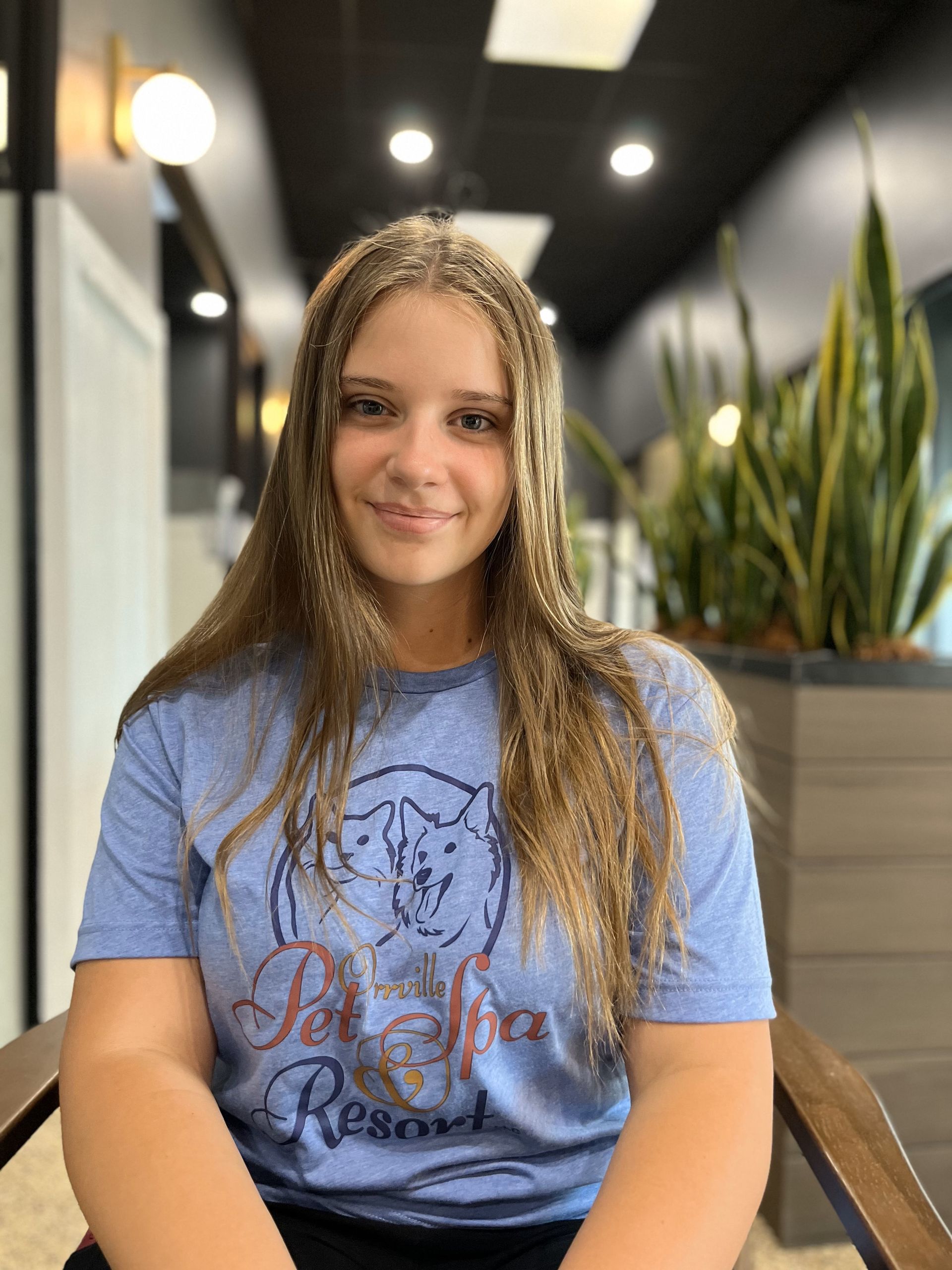 Young woman with long blonde hair sitting in a brown chair, wearing a blue t-shirt, smiling. She is in a modern interior with plants.