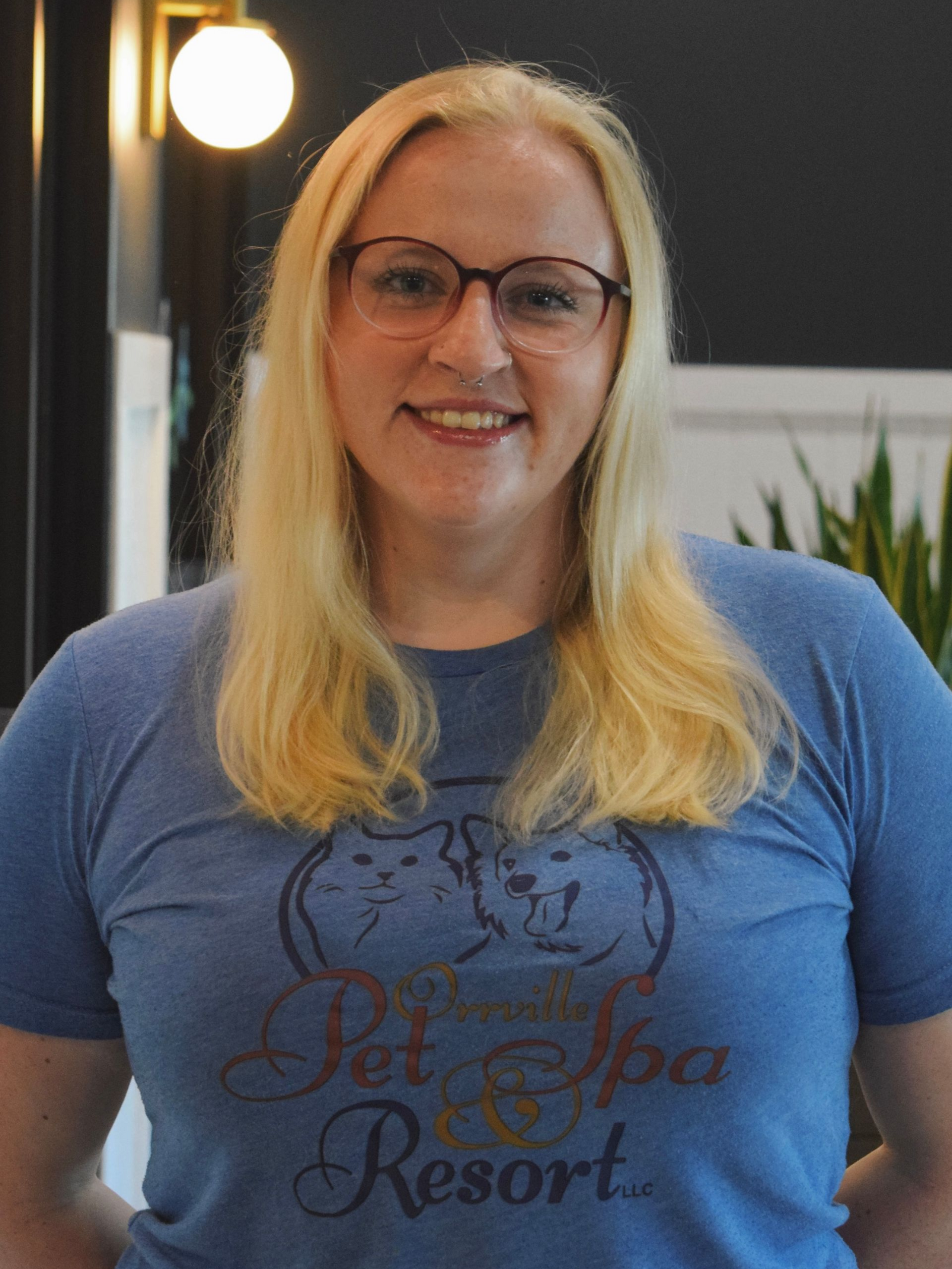 Young woman with blonde hair and glasses smiles at the camera. She wears a blue sweatshirt, set in a brightly lit interior.