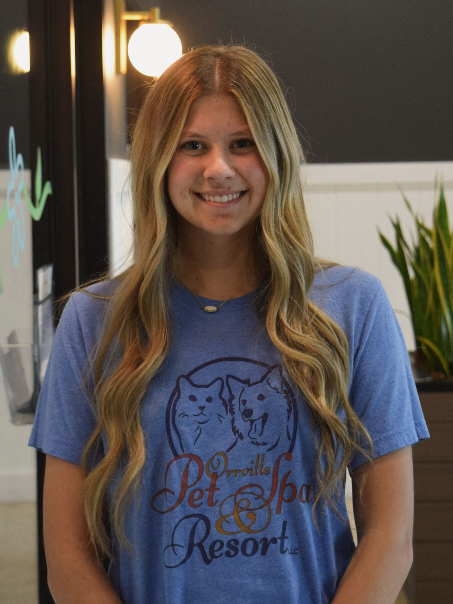 Young woman with long brown hair smiles, wearing a blue shirt, seated indoors.