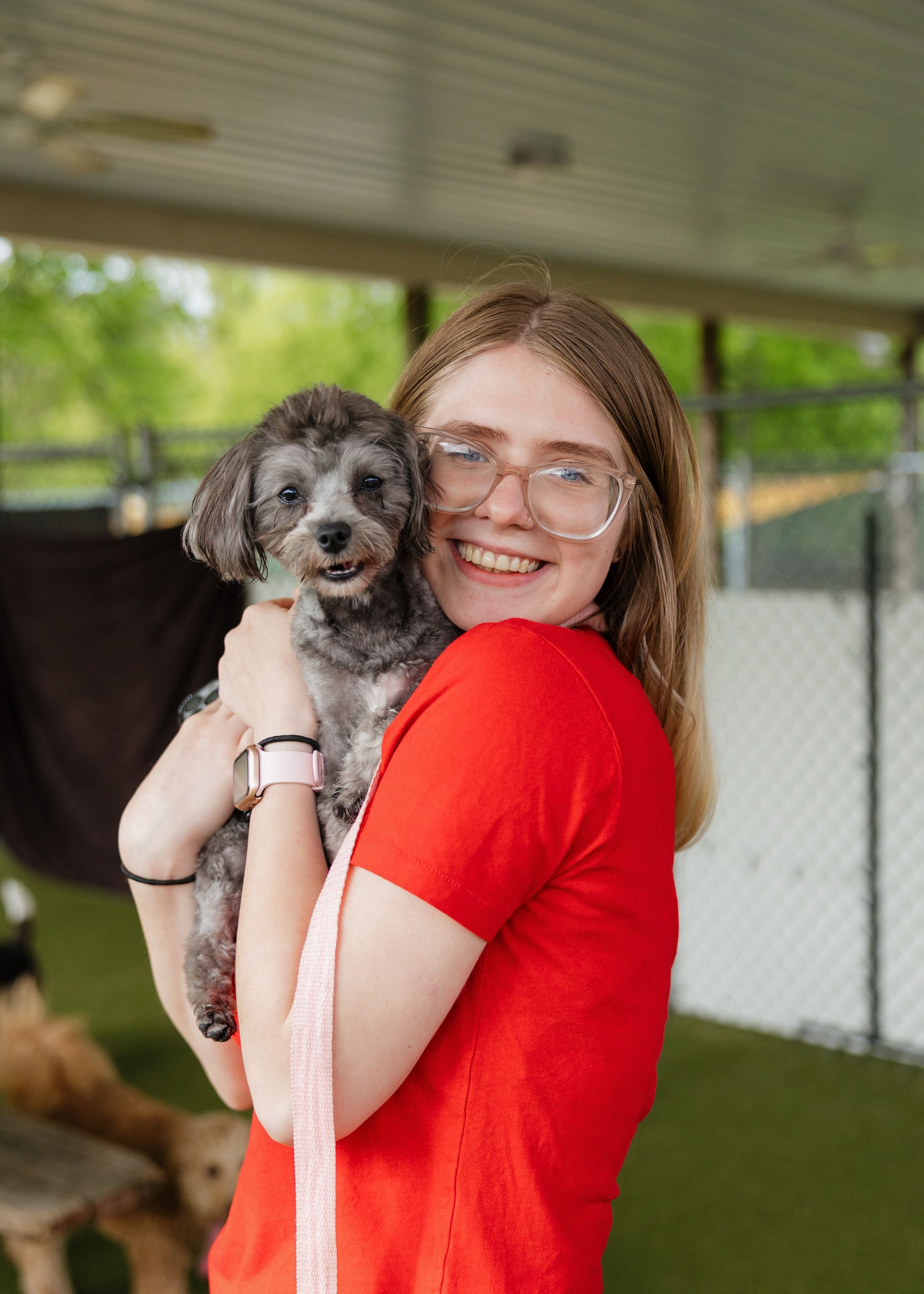 Woman with glasses in a red shirt smiles, holding a small, gray dog on a green surface, under a sheltered area.