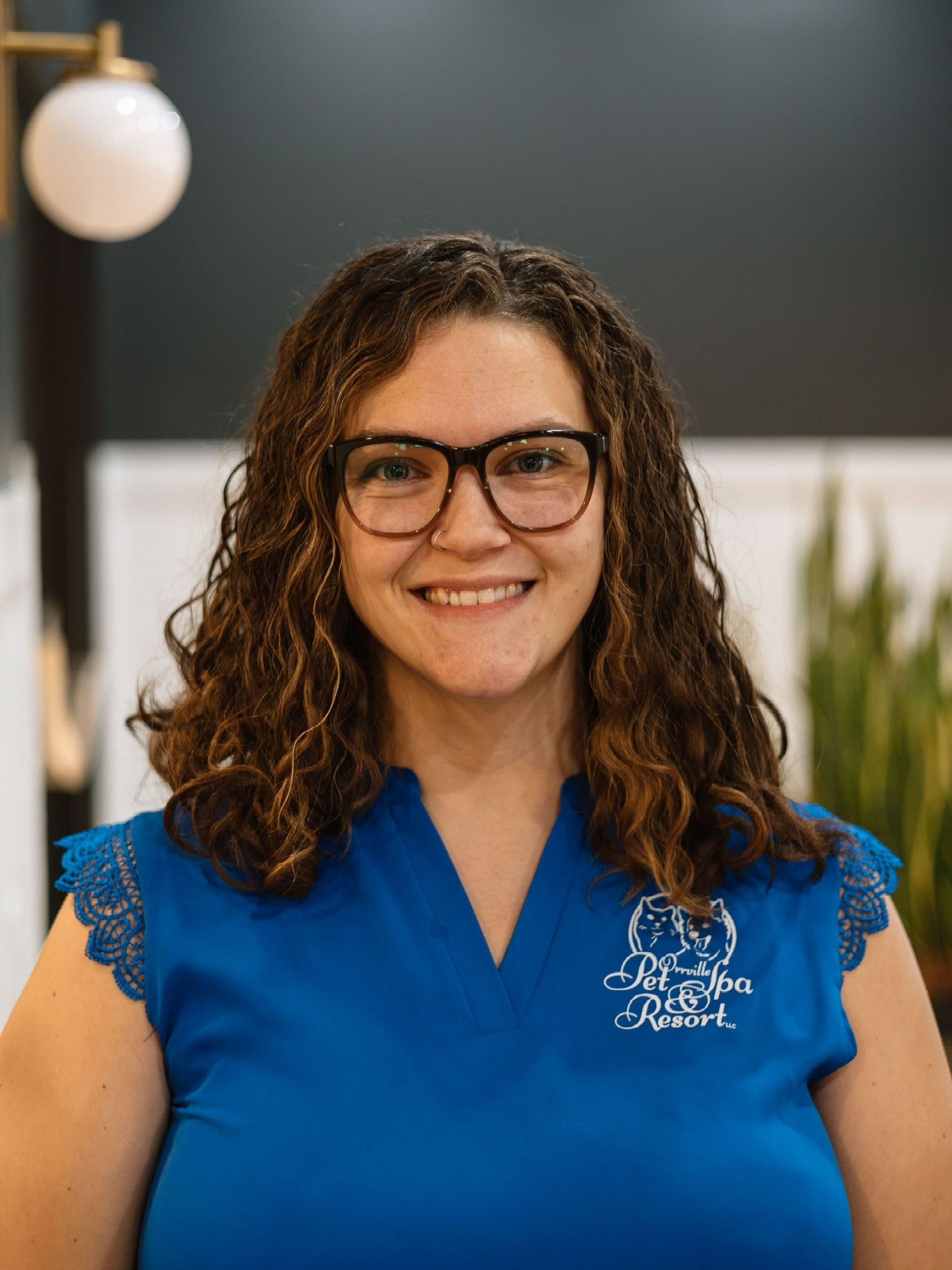 Woman with curly hair, glasses, and blue shirt smiles at the camera in an office setting.