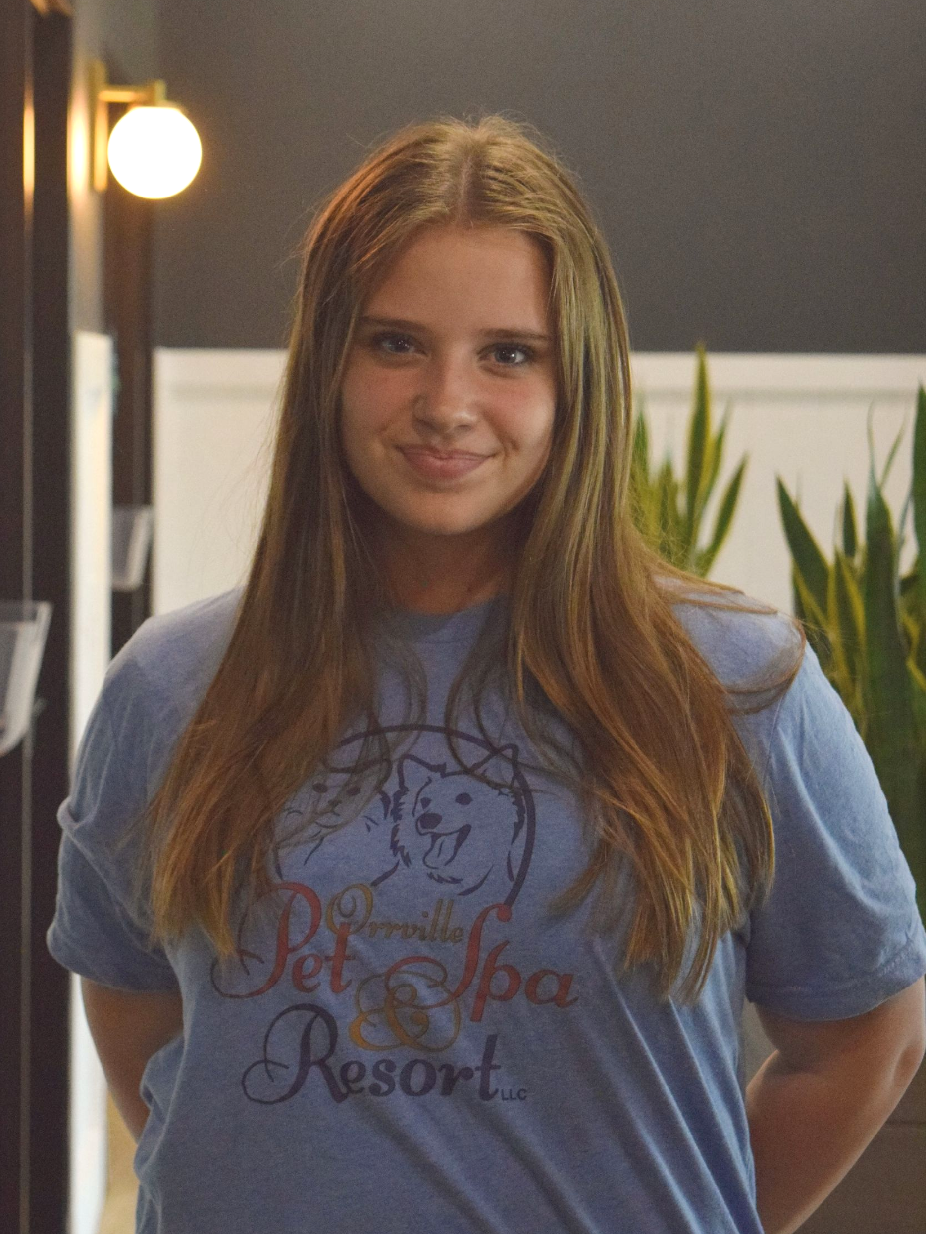 Young woman with long blonde hair sitting in a brown chair, wearing a blue t-shirt, smiling. She is in a modern interior with plants.