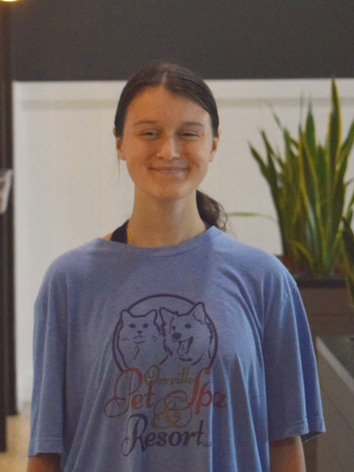 Woman with short brown hair smiles, wearing a blue t-shirt with a small logo, indoors.