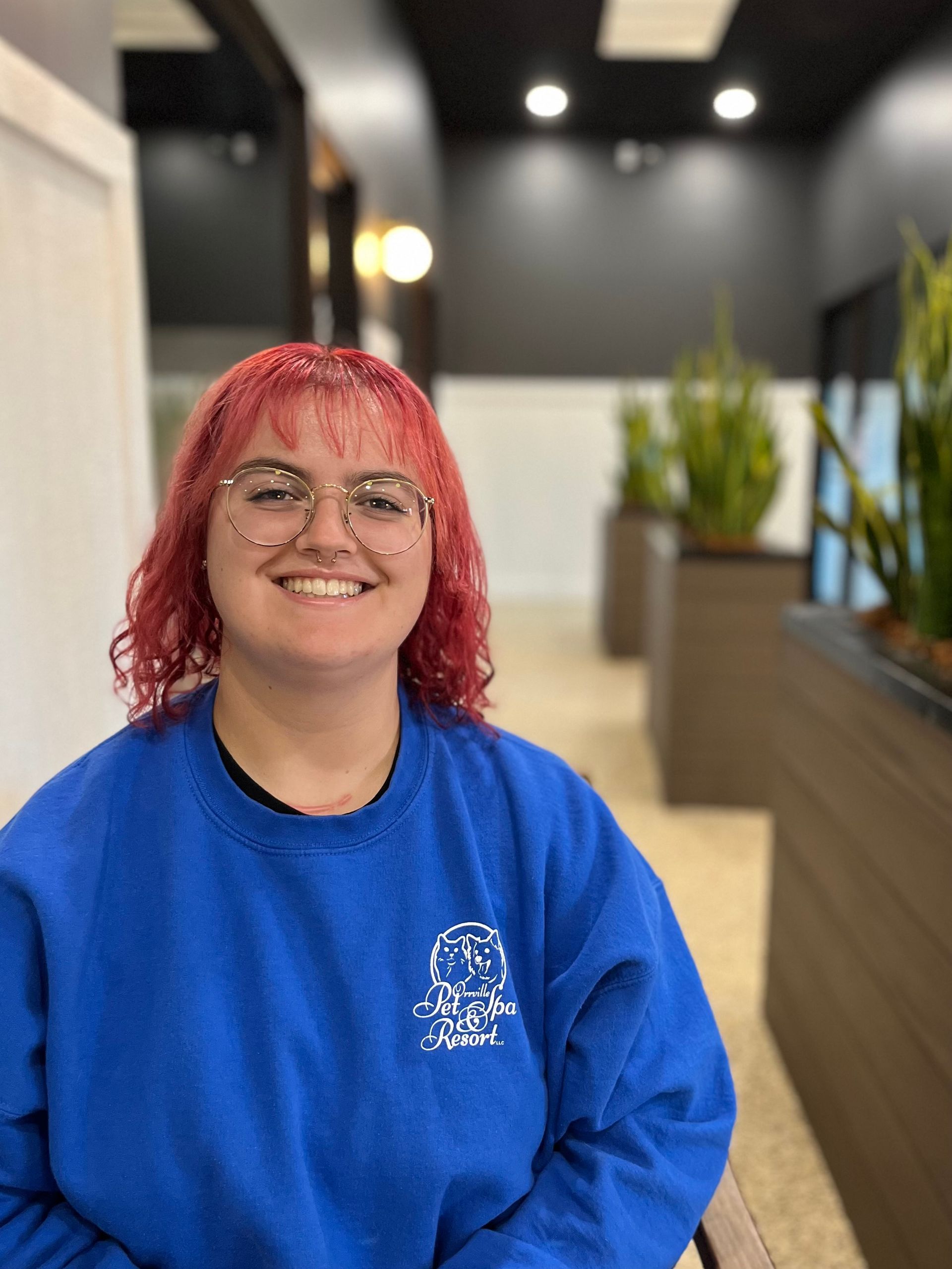 A young woman with red hair and glasses smiles at the camera, wearing a blue sweatshirt. She sits indoors near plants.