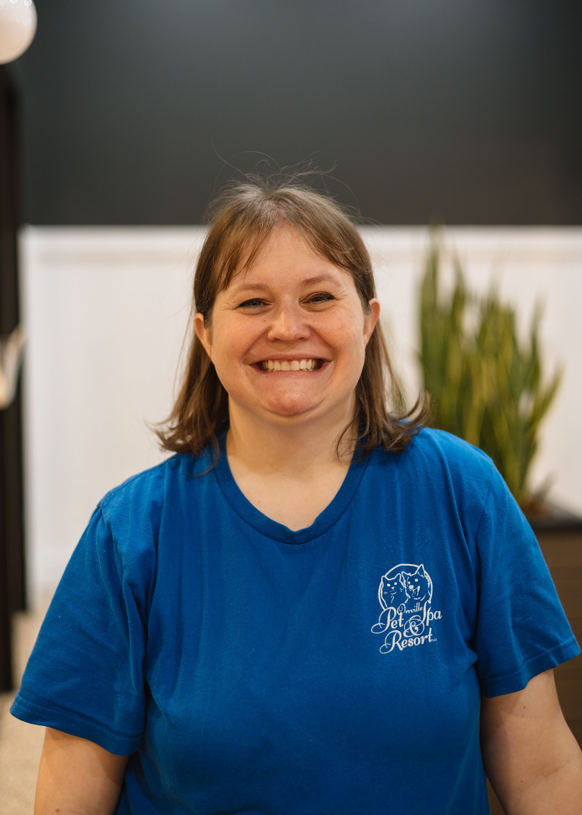 Smiling woman wearing a blue t-shirt. She has brown hair and stands indoors with a plant in the background.