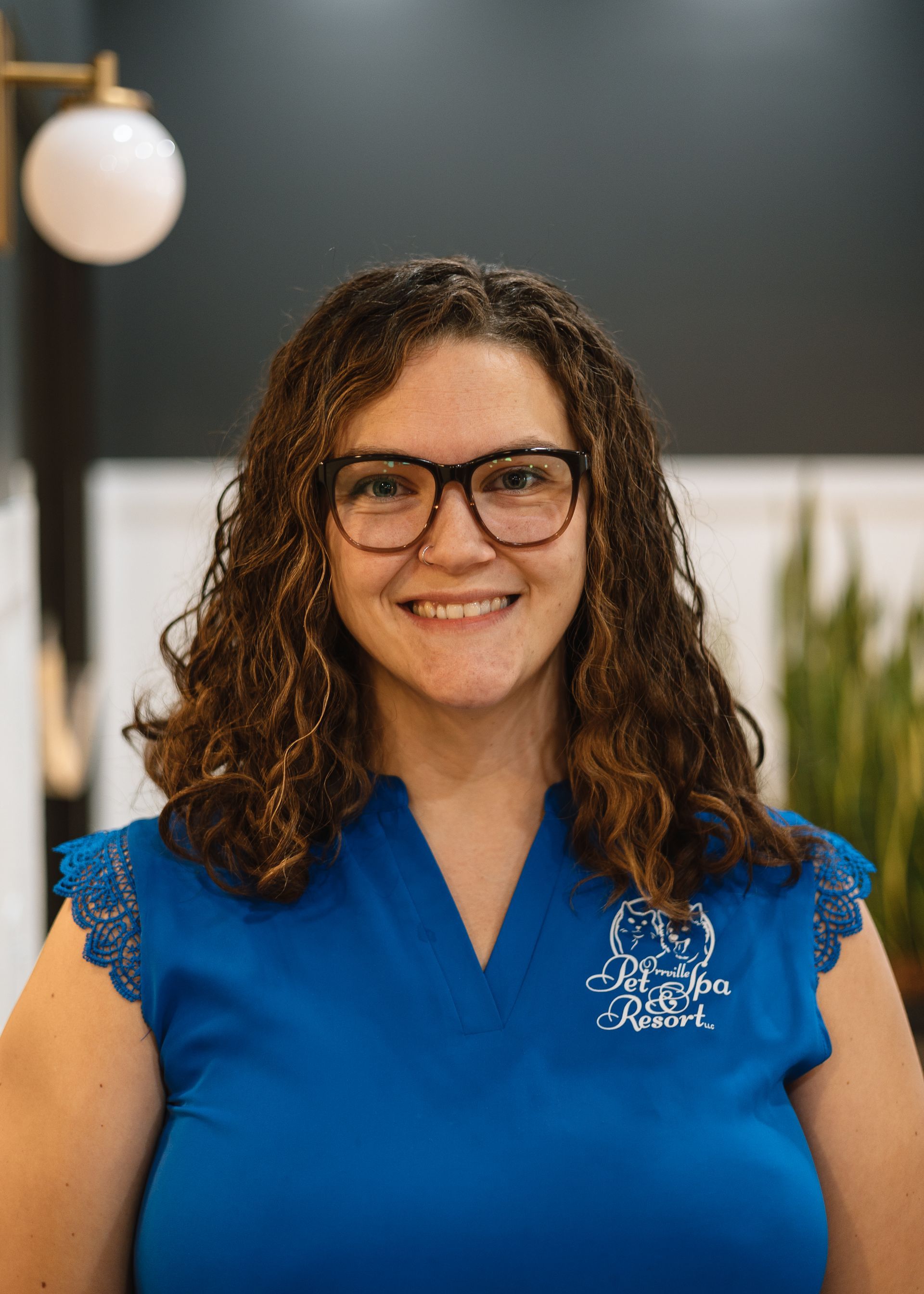 Woman with curly brown hair, wearing glasses and a blue shirt with the