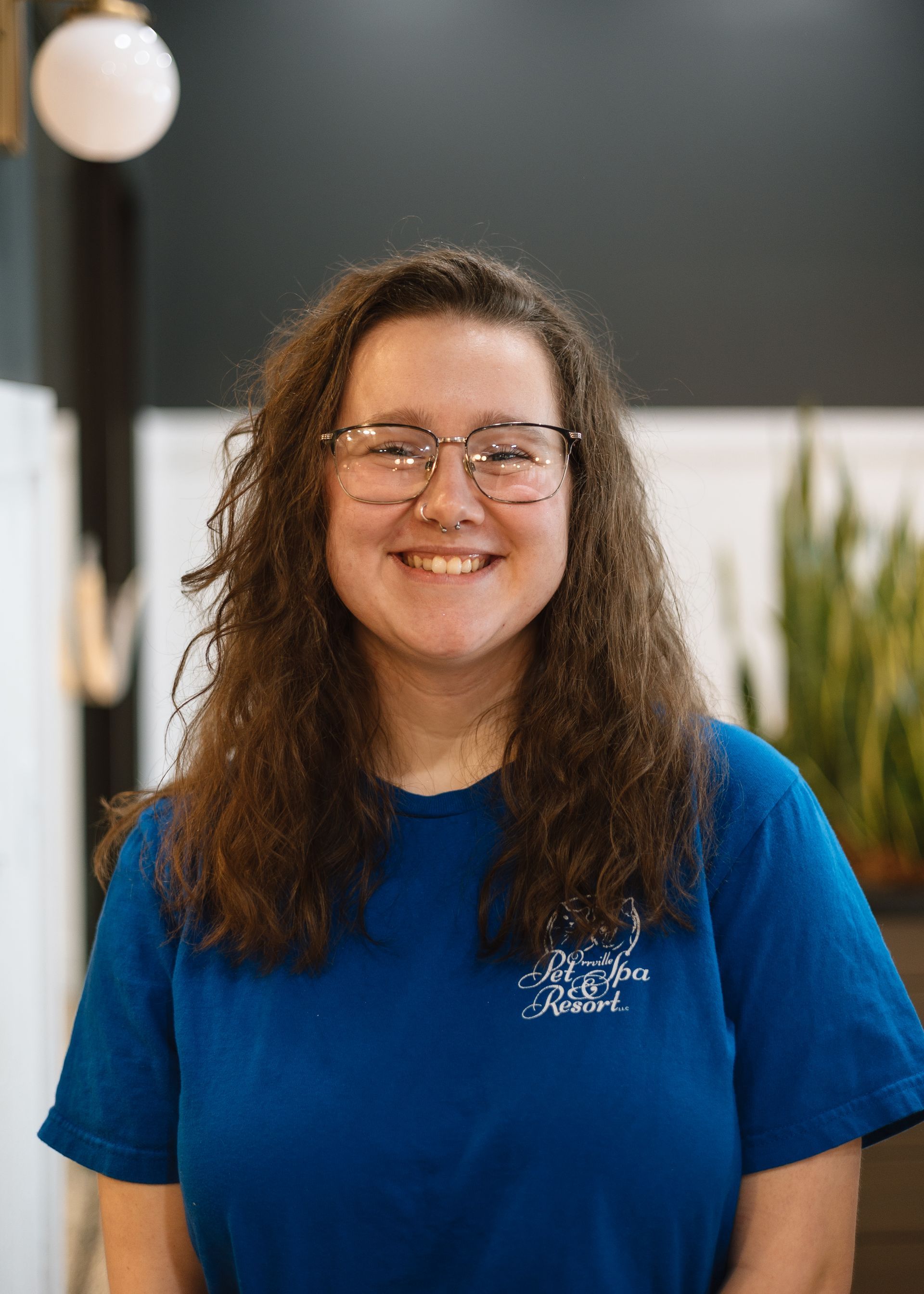 Smiling woman with glasses and curly hair wearing a blue shirt, inside a building with plants.