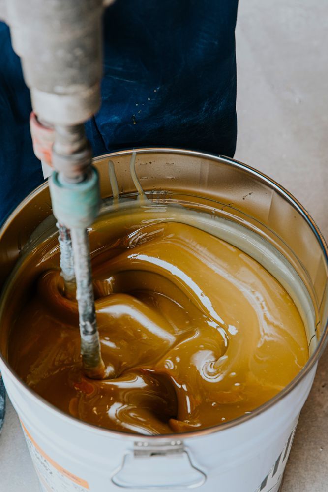 A person is mixing a yellow liquid in a bucket with a drill.