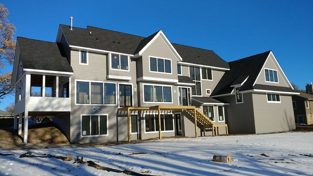 Gray two-story house with black roof, large windows, and wooden deck. Snow on the ground, clear blue sky.