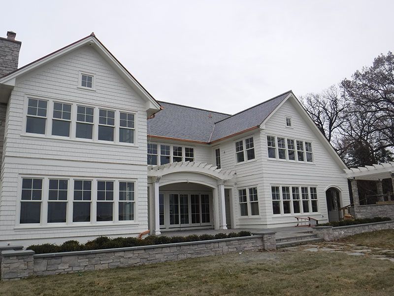 White two-story house with many windows and a stone wall at the base. Overcast day.