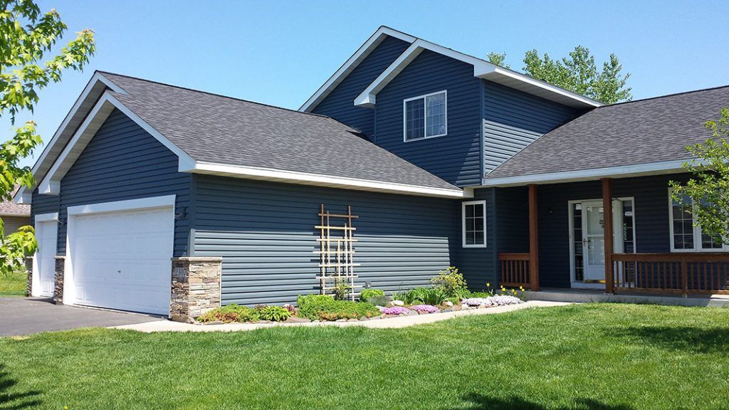 Blue-sided house with white garage doors, a small porch, and a green lawn on a sunny day.