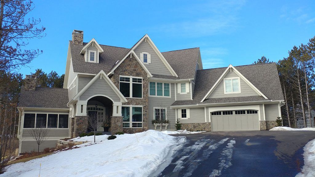 A large gray house with stone accents, a snow-covered driveway, and a blue sky.