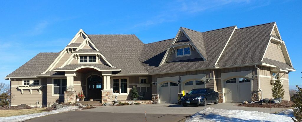 A large house with a gray roof and tan siding. A black car sits in the driveway. Clear blue sky.