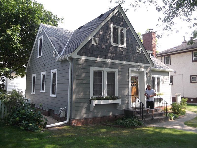 Gray house with a dark gray roof, a person on the porch, green lawn, trees.