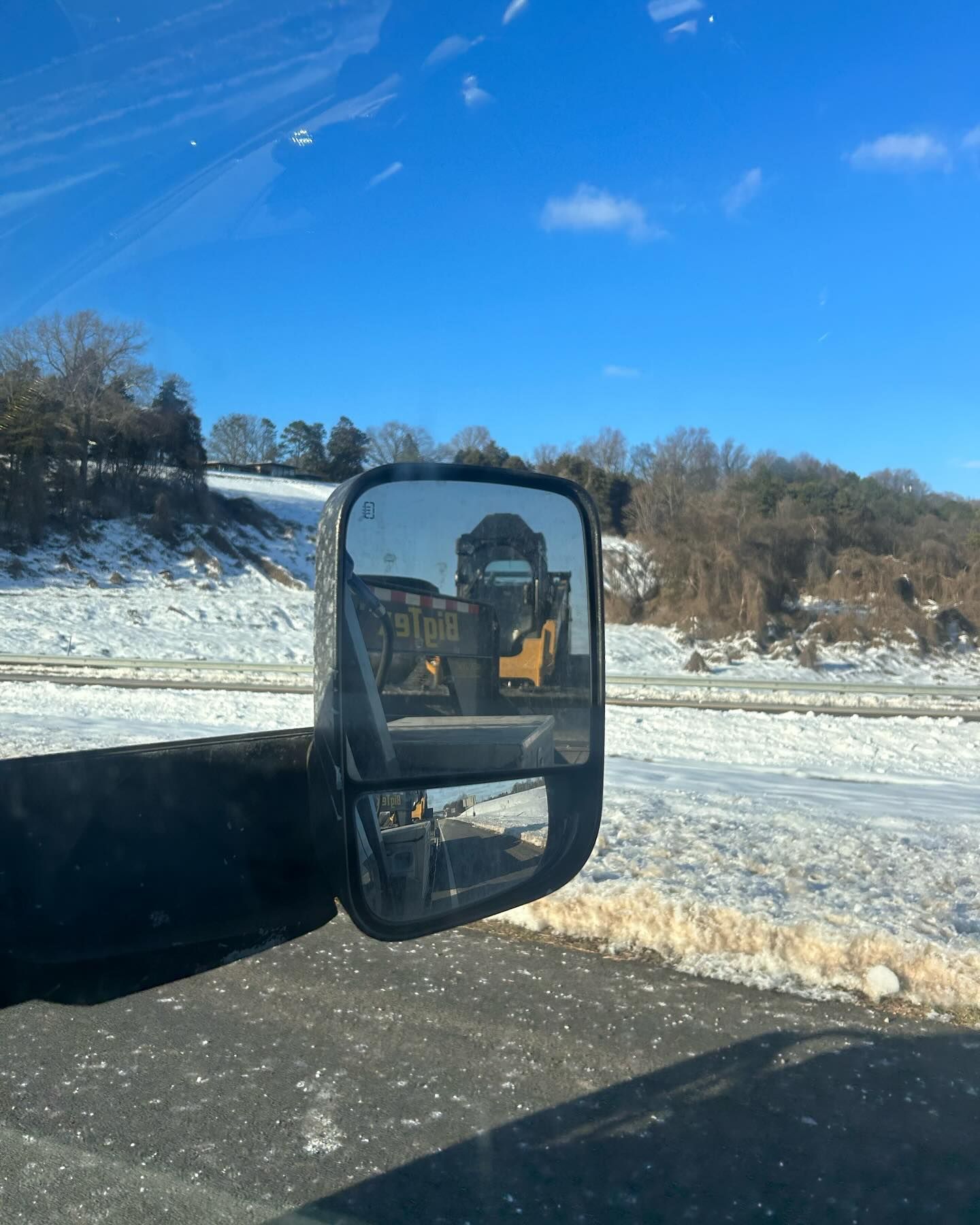 Side mirror reflecting a yellow construction vehicle on a snowy landscape under a blue sky.