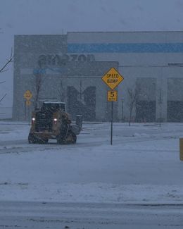 Snowy scene: a yellow front-end loader clearing snow in front of an Amazon warehouse. Sign with speed limit.