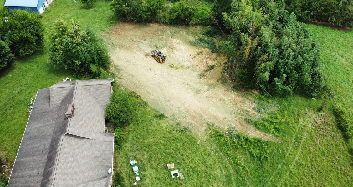 Aerial view of a clearing being worked on by a vehicle in a grassy area, near a house and trees.