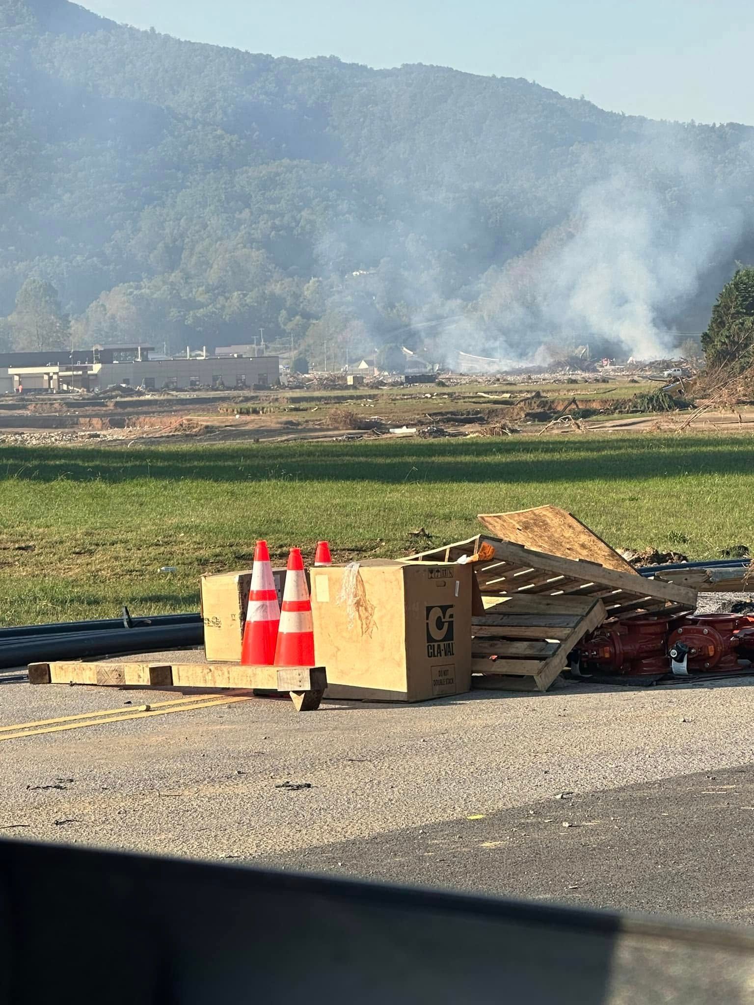Roadside view of smoke billowing from a fire in a field near a mountain range. Cones, pallets, and a box in the foreground.