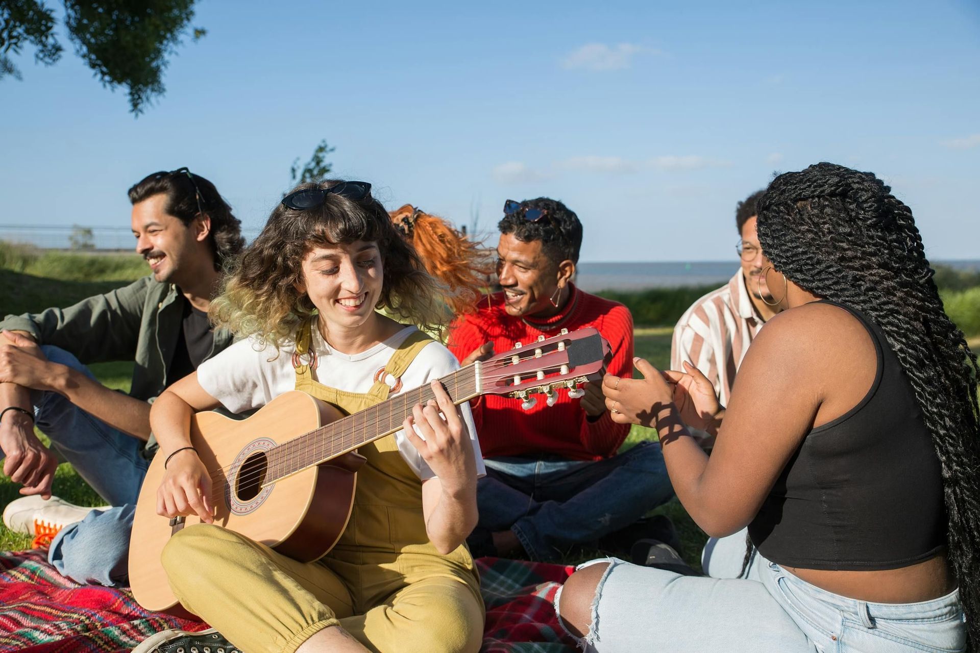 A group of people are sitting on a blanket in a park playing guitars.