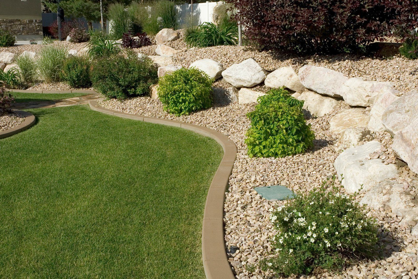 Green grass lawn with a curved border, surrounded by gravel and plants, and large rocks.
