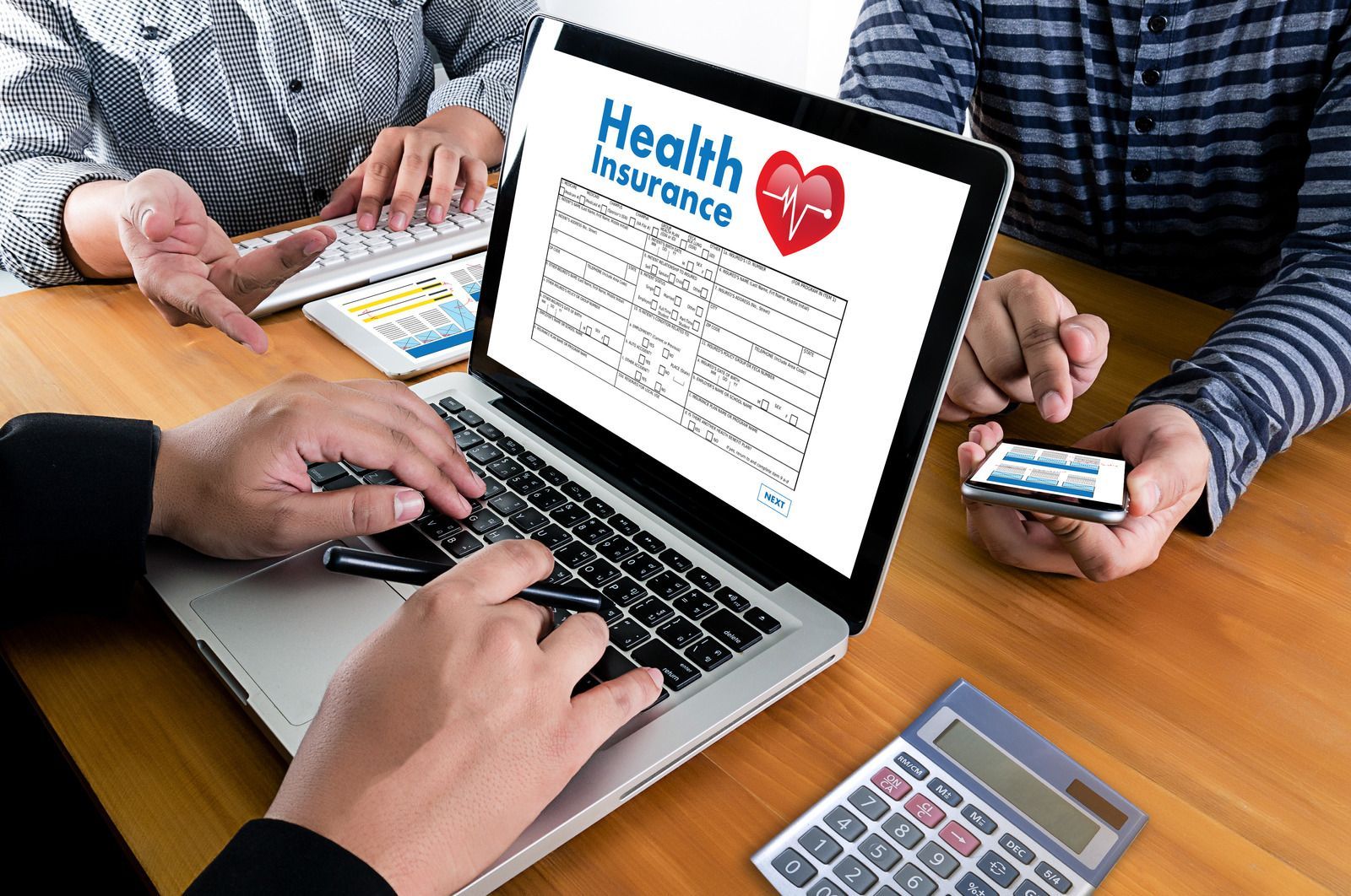 Hands around a laptop displaying health insurance, with documents and a smartphone on a desk