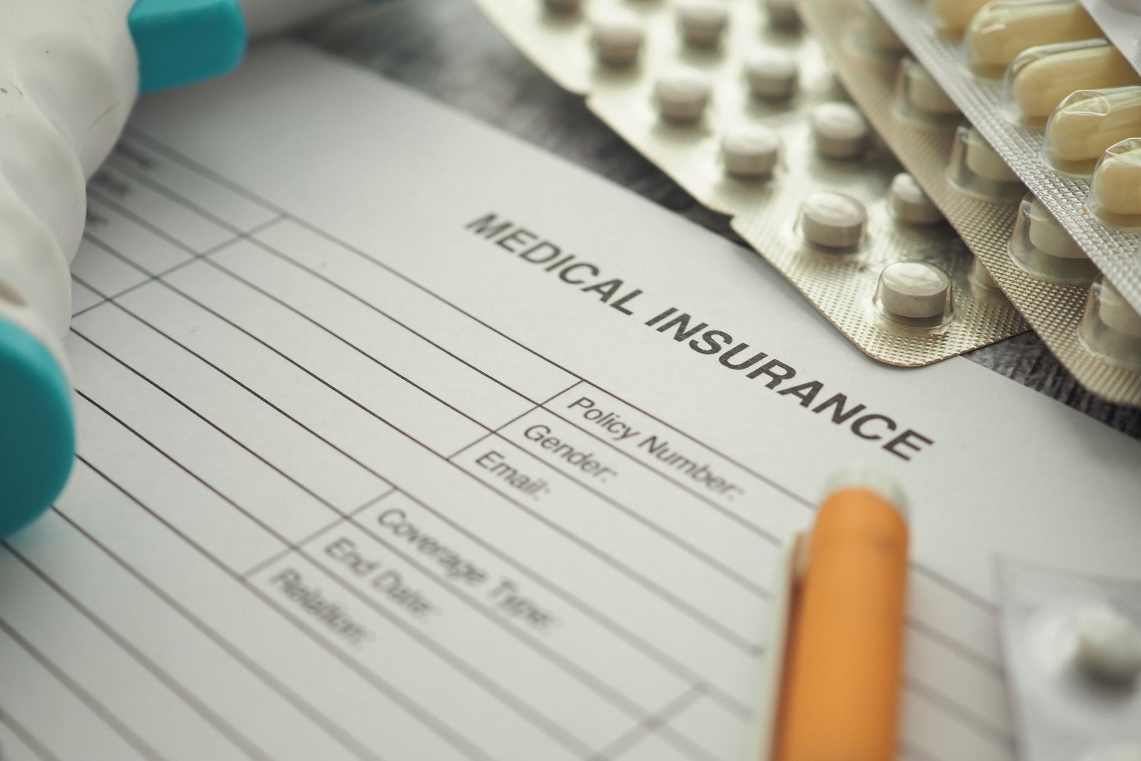 Medical insurance form with pill packs, teal gloves, and an orange pen on a desk