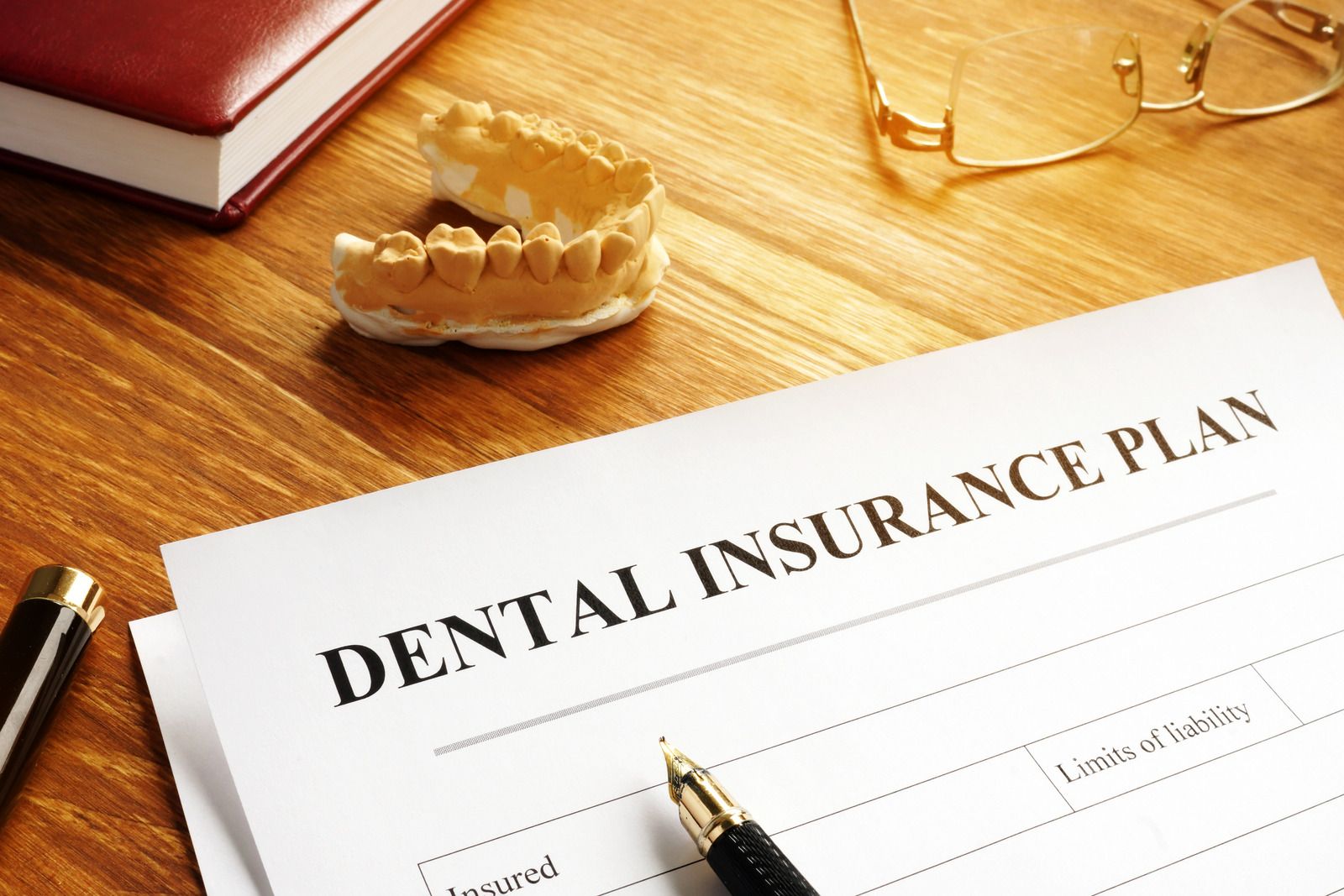 Dental insurance plan document on a wooden desk with a pen, glasses, and a book.