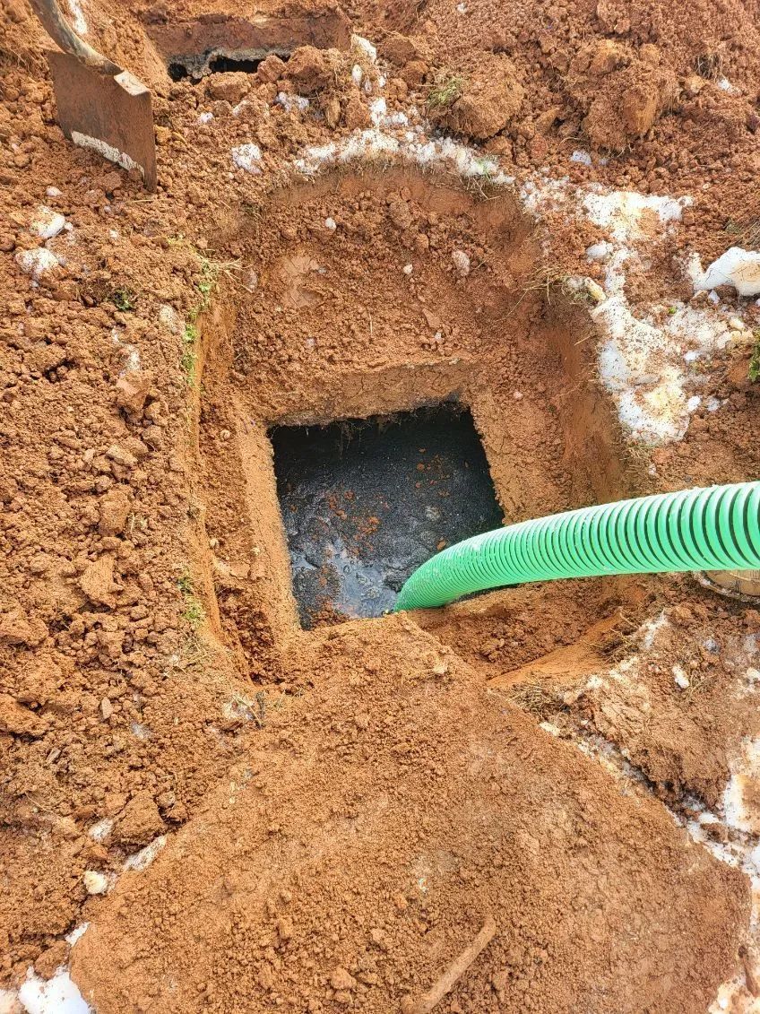 Green hose inserted into a square hole in reddish dirt or clay soil near a concrete edge