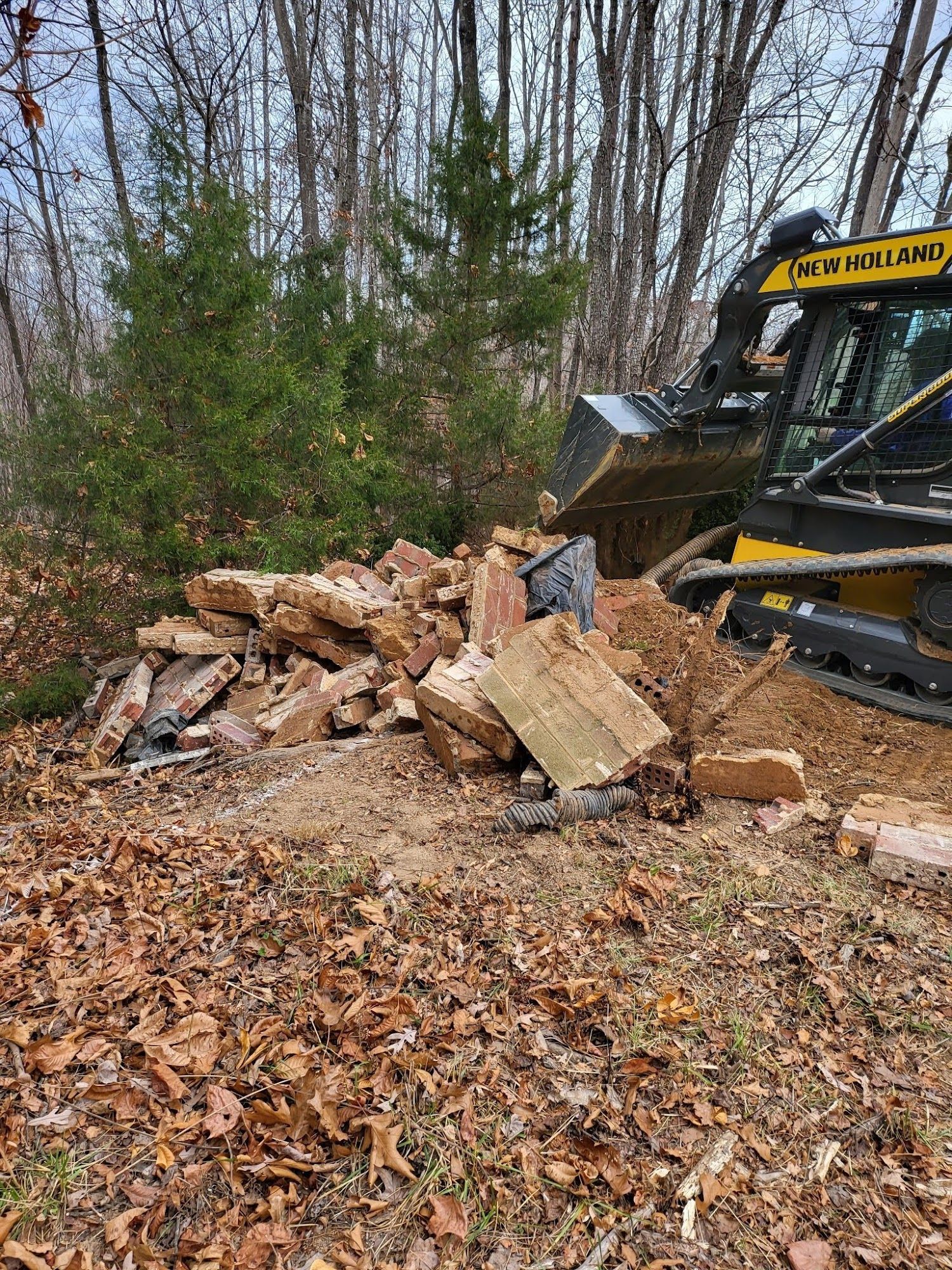 A New Holland skid steer moving a pile of rocks and debris near a wooded area.