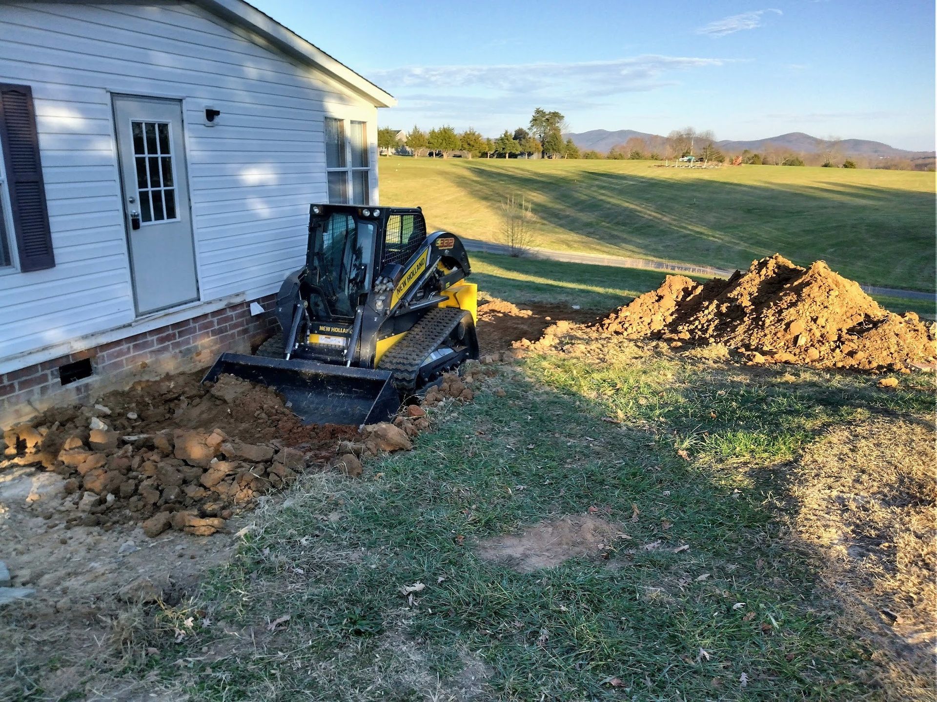 A small bulldozer digs beside a house, earth piled nearby. Green grass and a field are in the background.