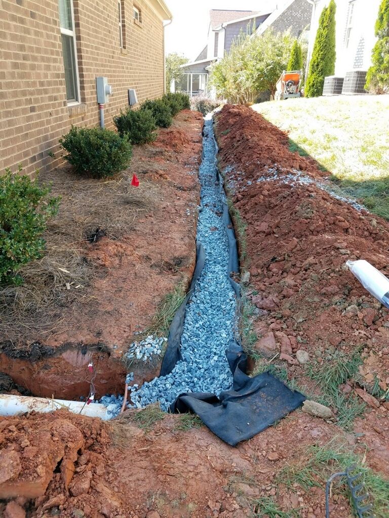 Trench filled with gravel, lined with fabric, for drainage along a brick building and grass lawn.