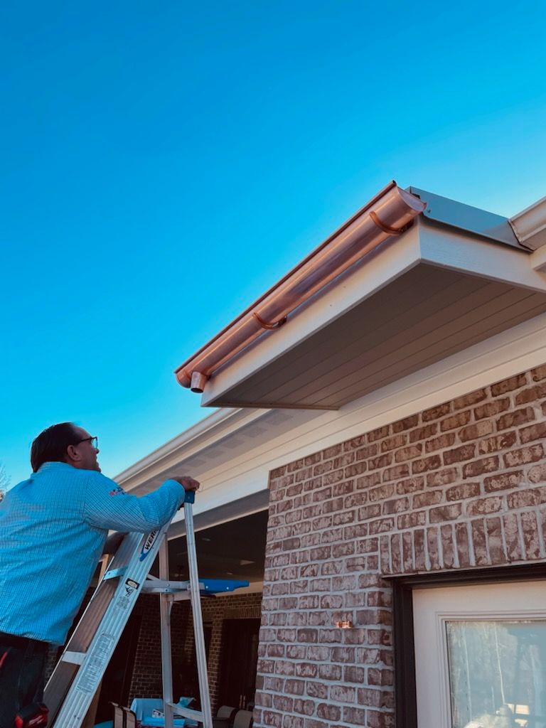 Man on a ladder installing copper rain gutter on a brick house with blue sky background.