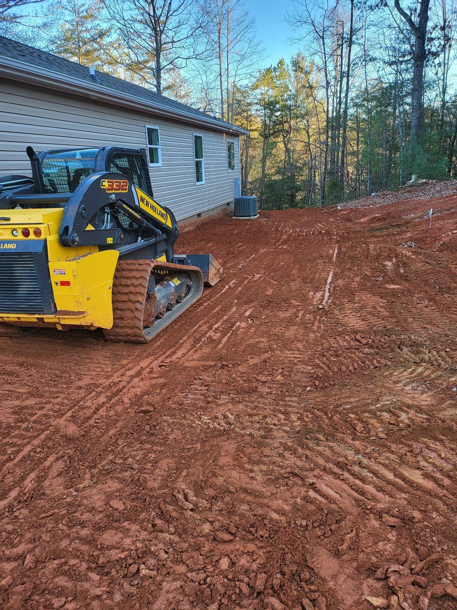Yellow skid steer on a red dirt slope next to a house with clear sky and trees in the background.