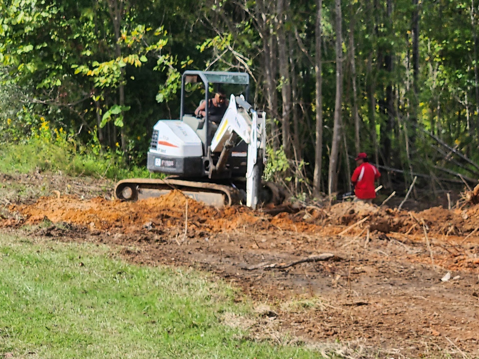 Bobcat excavator digging in dirt near trees; a person in red jacket watches.