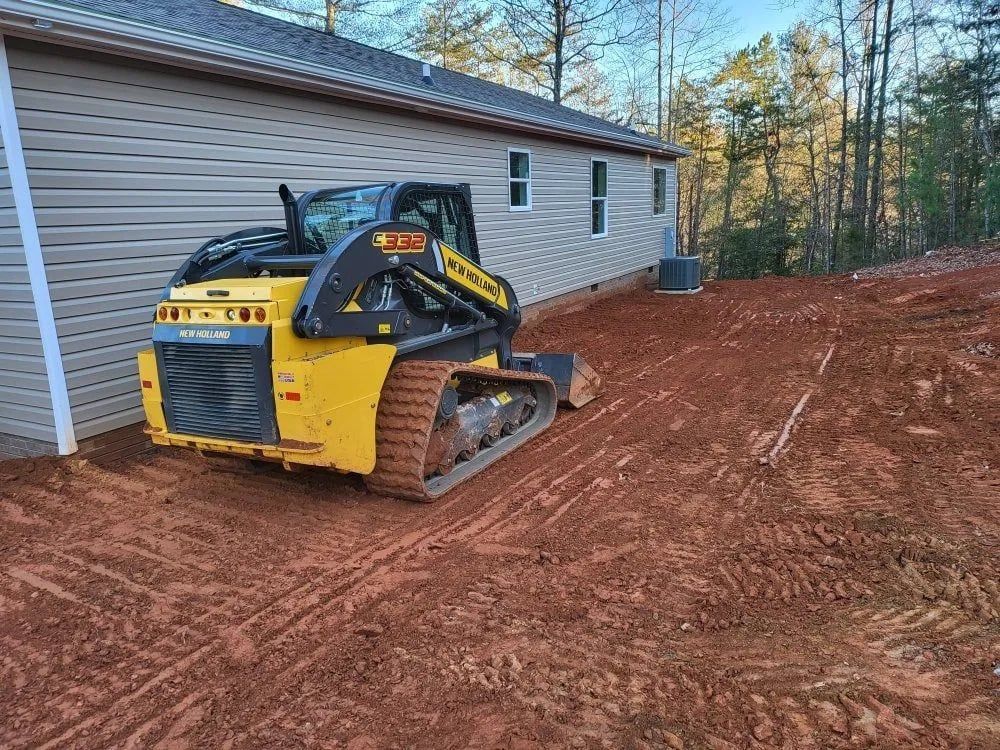 Yellow skid steer grading red dirt next to a beige house.