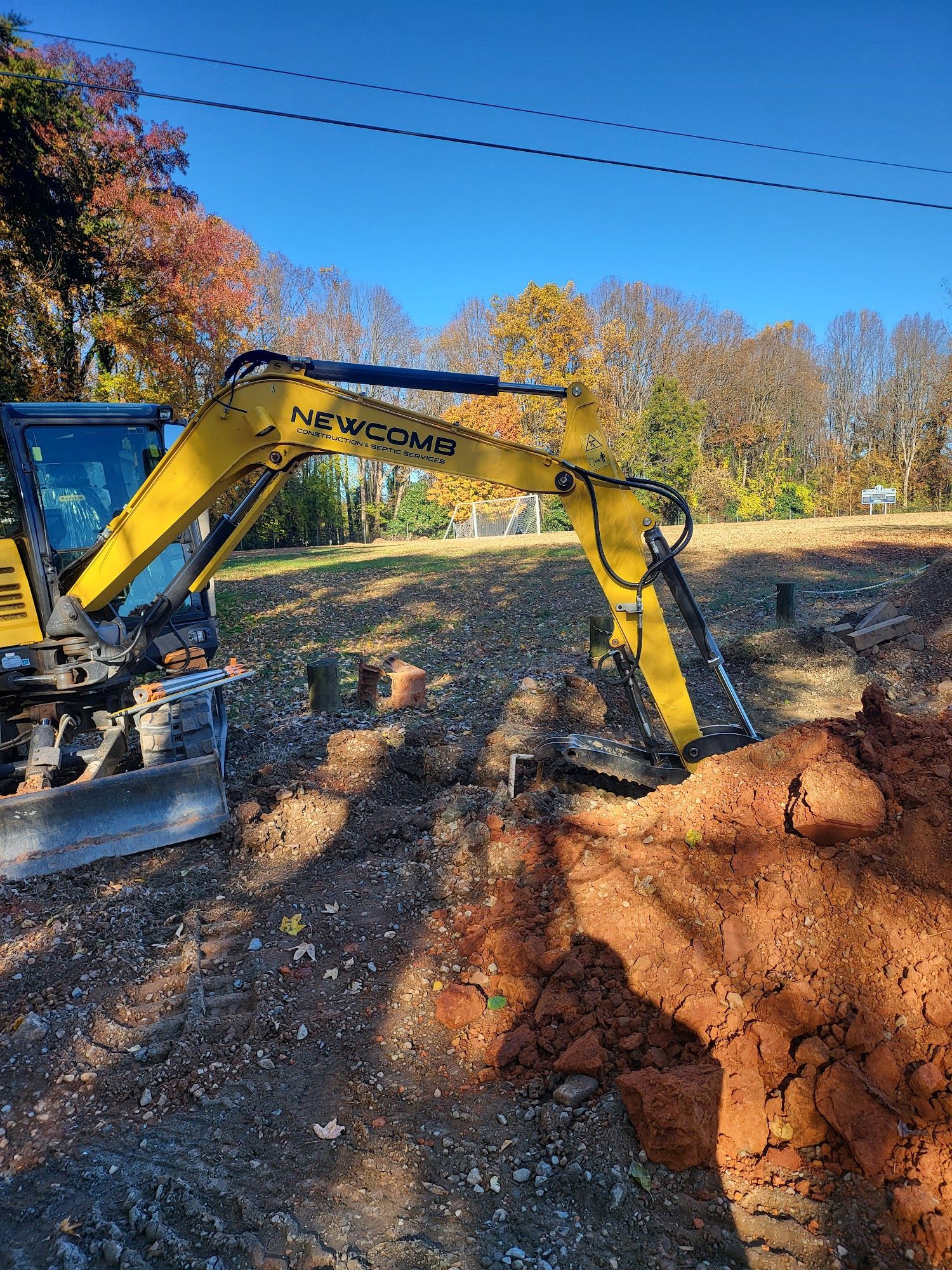 Yellow excavator digging in a dirt area, autumn trees and blue sky background.