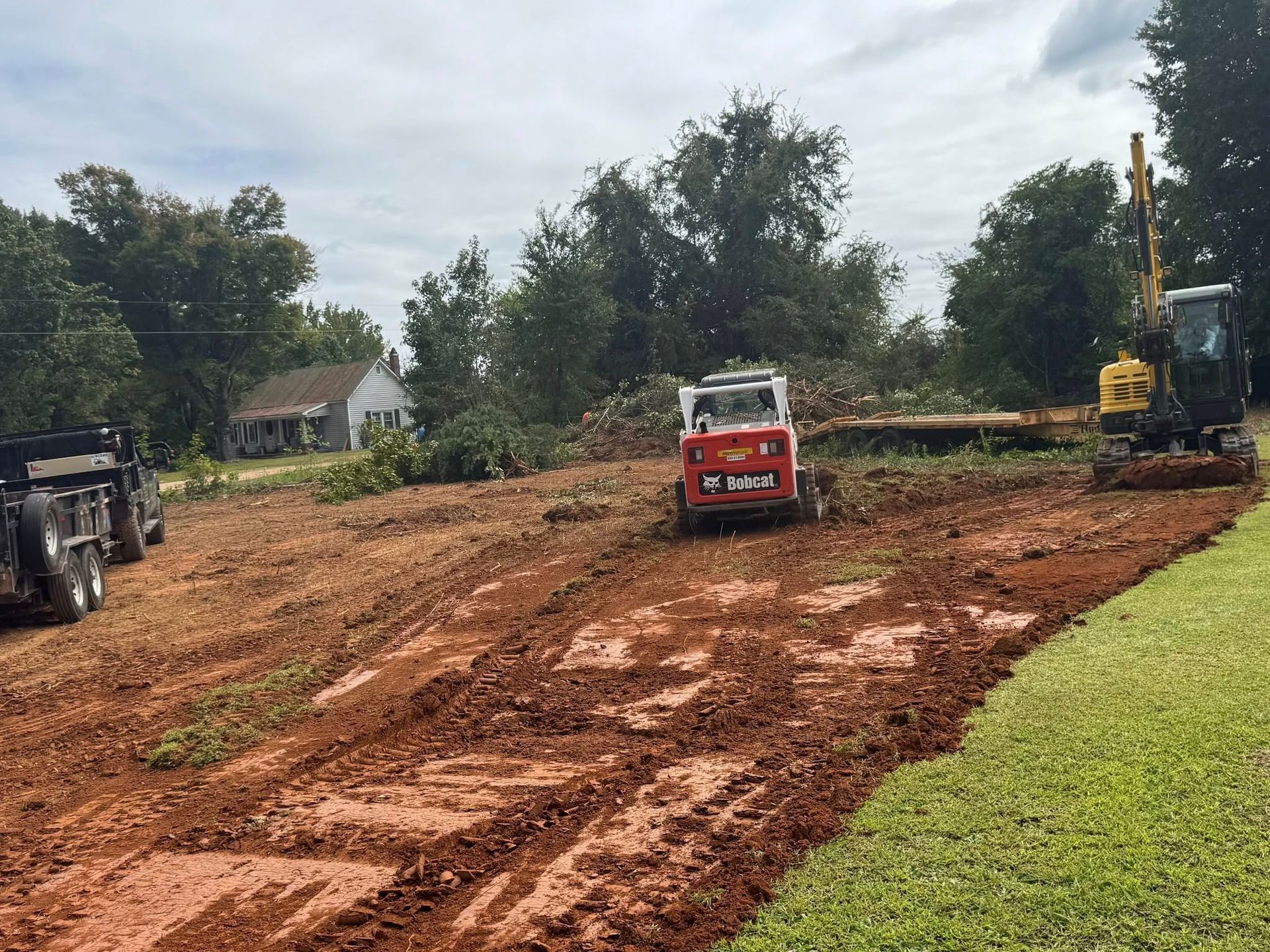 Construction site: Skid steer and excavator working on graded, muddy land with a trailer, trees, and house in background.
