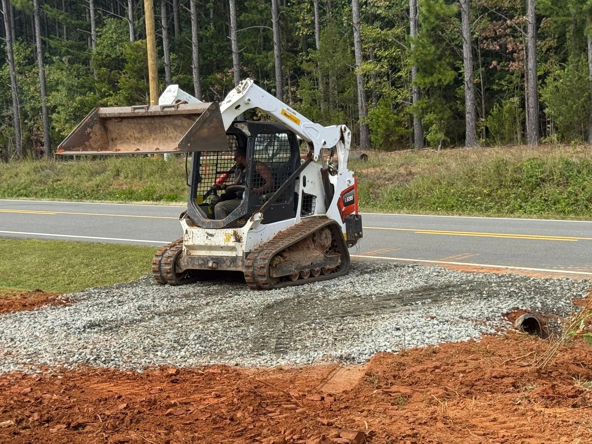 A white Bobcat skid-steer loader on gravel, near a road and woods.