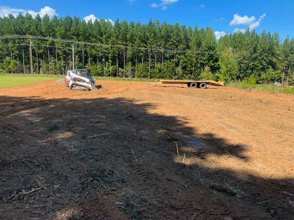 A small skid steer tractor on a cleared, brown dirt field, trailer, trees, and blue sky.