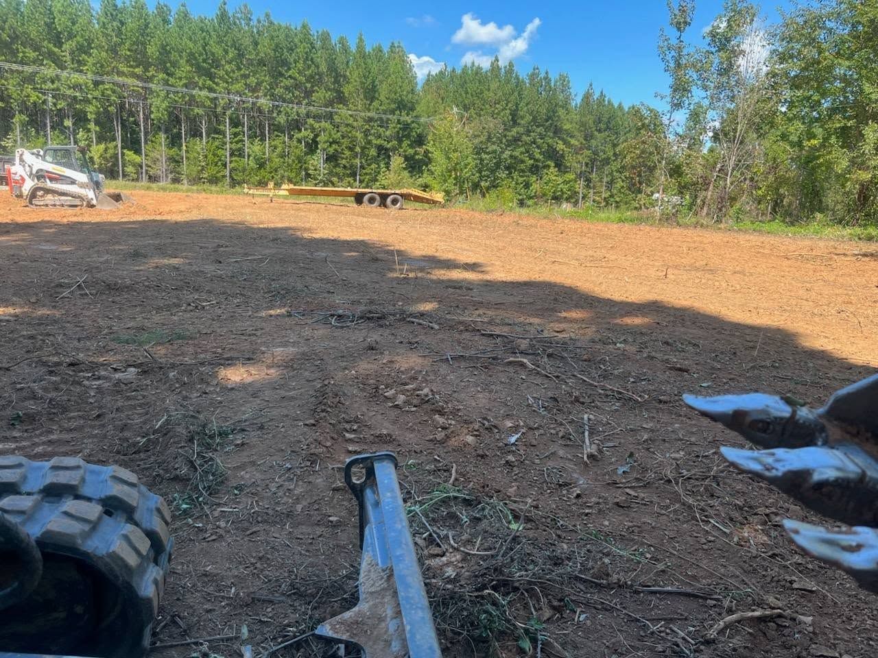 Clearing a dirt lot, with equipment in the foreground and a forest in the background. Blue sky overhead.