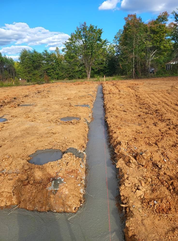 A freshly poured concrete foundation trench in a dirt field under a bright sky with trees in the background.
