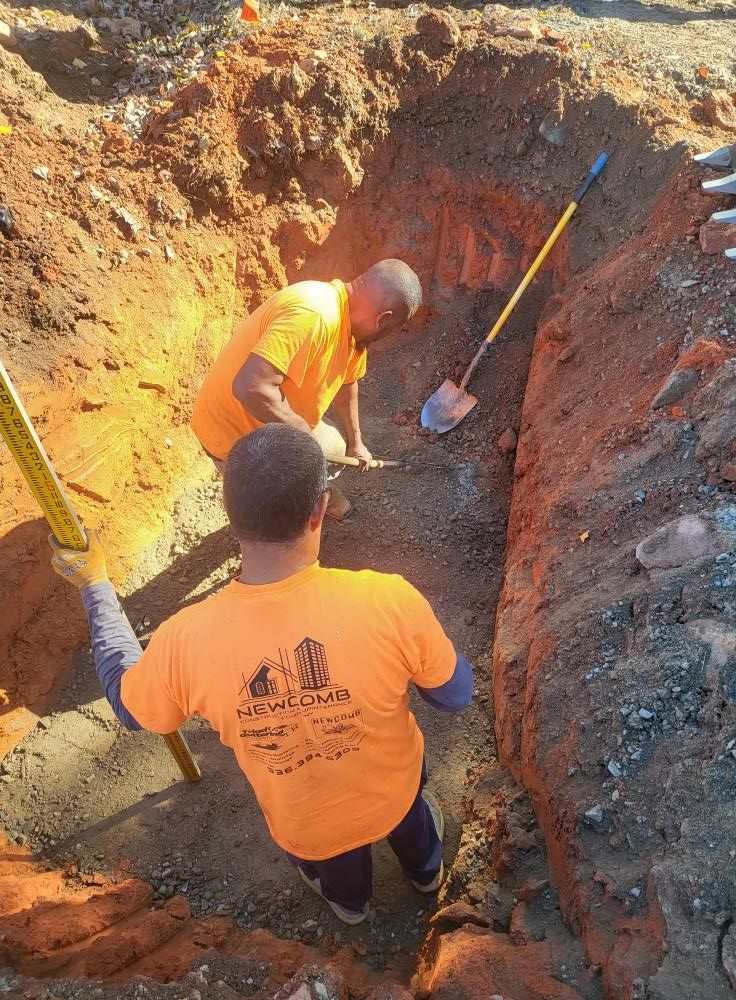 Two workers in orange shirts excavating a trench, one using a shovel, one measuring with a level.