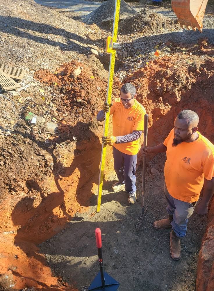 Two construction workers in orange shirts using a level inside an excavated trench.