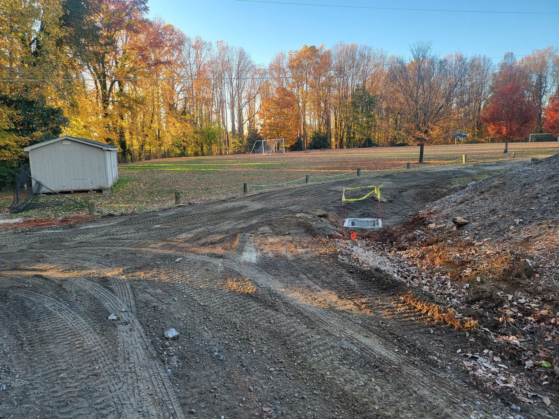 Construction site with dirt piles and small white shed, trees in background, autumn colors.