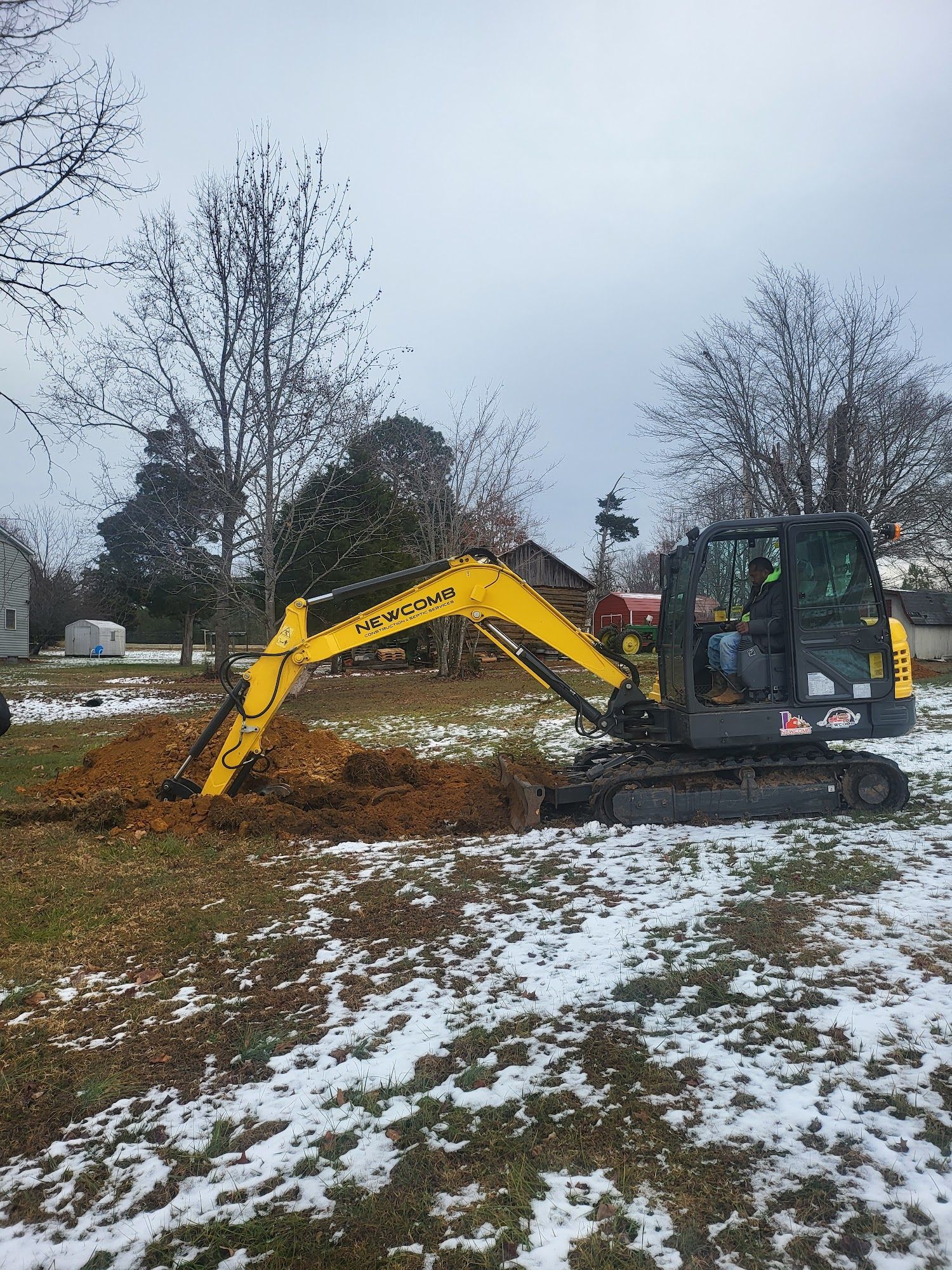 Yellow excavator digging in a snow-covered yard on an overcast day.