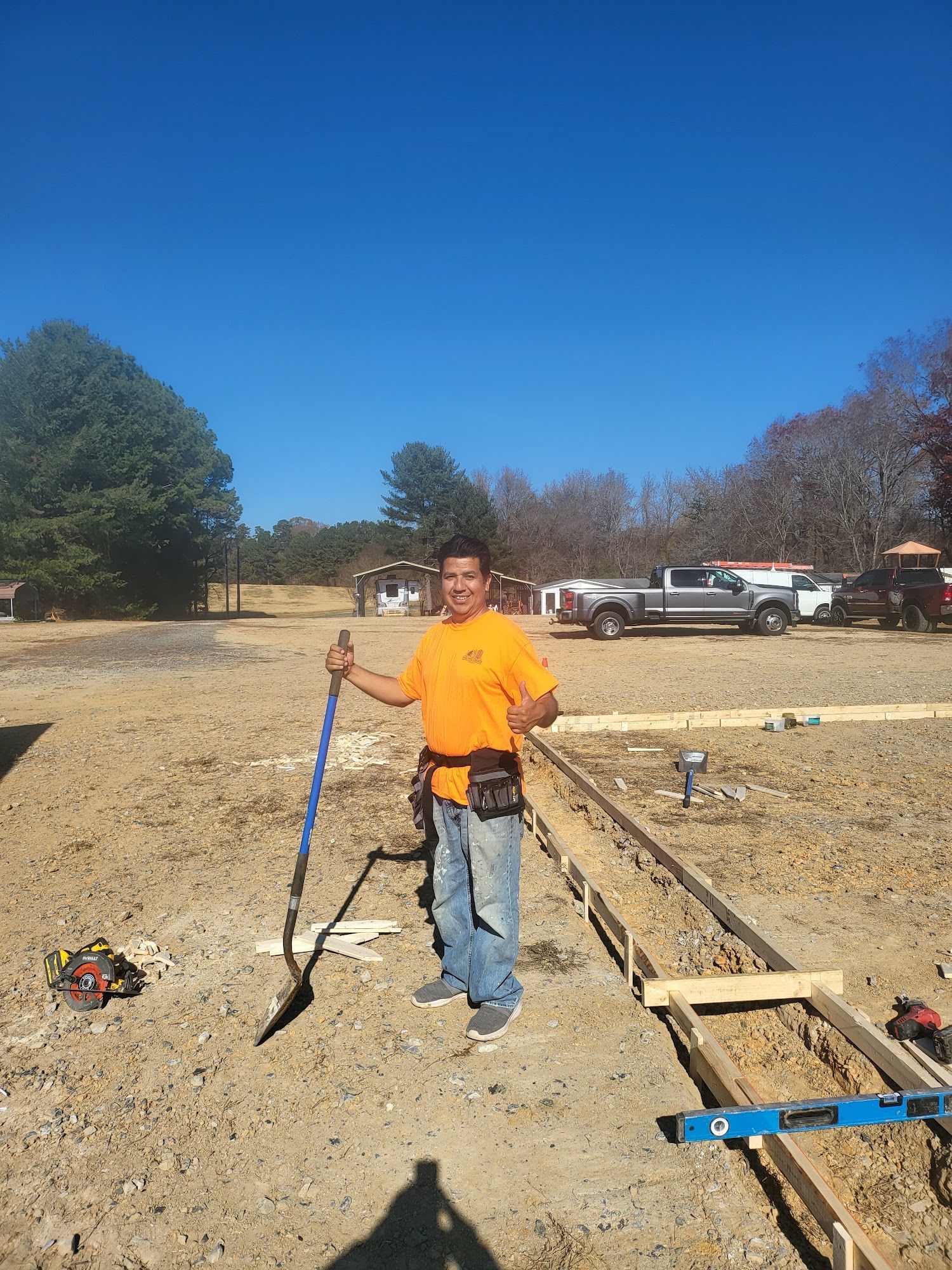 Man in an orange shirt holds a shovel on a construction site; clear blue sky.