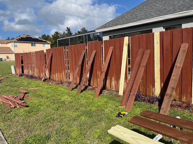 A damaged brown wooden fence being repaired outdoors, with replacement boards scattered on the grass.