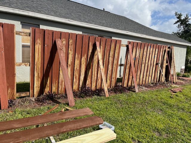 A damaged wooden fence along a house needs repair. The boards are weathered and missing.