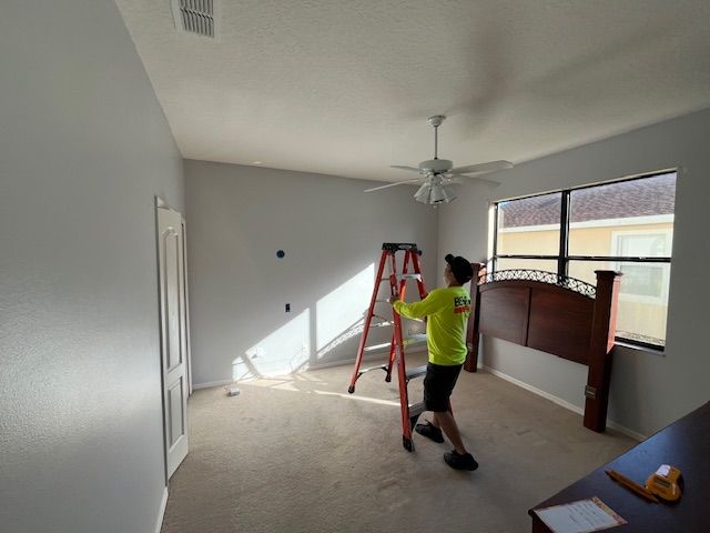 Person in a neon yellow shirt on a ladder, painting a light gray bedroom, with a headboard nearby.